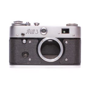 Op de afbeelding: Een vintage FED 3 camera met een zilveren bovenkant en een zwarte, getextureerde behuizing. De camera heeft verschillende draaiknoppen en een lensopening. De tekst "FED 3" staat op de voorkant. Op een witte achtergrond.