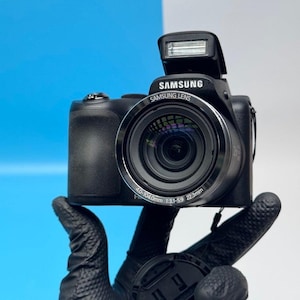 Pode incluir: Uma câmara digital Samsung preta com flash pop-up, uma lente Samsung com as especificações 4.0-104.0mm 1:3.1-5.9 22.3mm impressas no anel frontal e uma área de aderência texturizada na lateral do corpo da câmara.