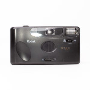Puede incluir: Cámara Kodak Star 320 negra. La cámara tiene flash, lente y botón disparador. La palabra "Kodak" está impresa en la parte delantera. La palabra "STAR" está impresa en la parte delantera de la cámara.
