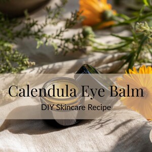 Może przedstawiać: Słoiczek balsamu do oczu z nagietka z pokrywką, umieszczony na neutralnej tkaninie. Tekst "Calendula Eye Balm" i "DIY Skincare Recipe" są nałożone na obraz. Żółte kwiaty nagietka i zieleń otaczają słoiczek.