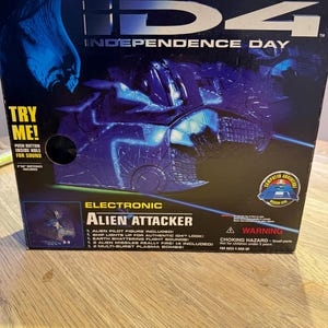 May include: A boxed electronic Alien Attacker toy from the film Independence Day. The box is mainly blue and black, with the film's title and a spaceship graphic. The box includes the text "Try Me!" and a warning about small parts.