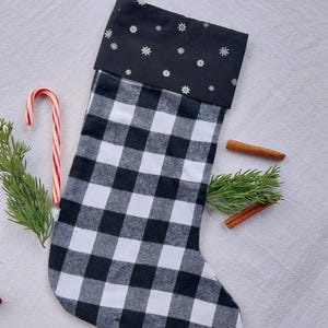 May include: A Christmas stocking featuring a black and white buffalo check pattern on the main body and a black cuff decorated with small white snowflake icons. The stocking includes a black fabric hanging loop at the top corner.