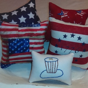 May include: Four decorative throw pillows with patriotic themes. The pillows feature red, white, and blue colors with stars and stripes patterns. One pillow has the year 1776 embroidered on it. Another pillow has a simple embroidered design of a top hat.