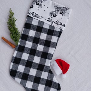 May include: A Christmas stocking featuring a black and white buffalo check pattern on the body and a white cuff with black line art of pine branches, bells, and stars, with the text Merry Christmas printed twice across the cuff.