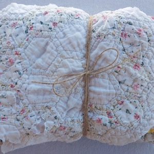 May include: A folded stack of quilted fabric pieces tied together with thin brown twine. The top layer features a white background with a repeating floral pattern of small pink and green flowers, while other layers show yellow and green fabric.