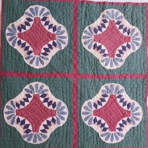 May include: A quilted textile featuring four repeating floral-like patterns. Each pattern has a central burgundy diamond shape, surrounded by blue and white details, set against a green background. The quilt is stitched with a grid pattern.