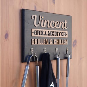 Könnte beinhalten: Schwarzes Holzschild mit dem Namen "Vincent" in hellbrauner Schrift. Darunter steht "GRILLMEISTER" und "GRILLEN & CHILLEN" in ähnlicher Schrift. Das Schild hat Metallhaken, an denen Grillwerkzeuge und eine Schürze hängen.
