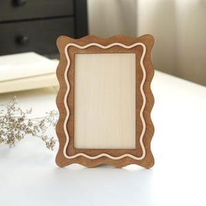 Op de afbeelding: Een houten fotolijst met een golvend randontwerp. De lijst is gemaakt van lichtbruin hout met een lichtere houten accent. De lijst is leeg, klaar om een foto of kunstwerk te bevatten. De lijst staat op een wit oppervlak.