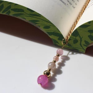 May include: A gold-toned metal chain bookmark featuring a dangling charm composed of a series of beads in varying shades of pink and translucent white, finished with a larger, vibrant magenta bead at the bottom, resting inside an open book.
