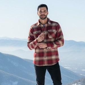 May include: A red and beige plaid flannel shirt with two chest pockets. The shirt is buttoned and worn with black pants. The background features a mountain range under a clear sky.
