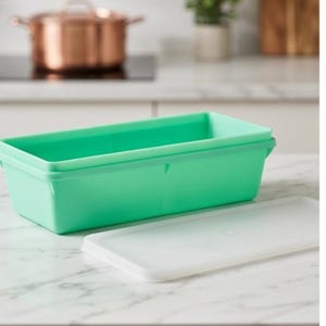 May include: A rectangular, mint-green plastic food storage container with a white lid. The container is empty and sits on a white marble countertop. A copper-colored pot is visible in the background.