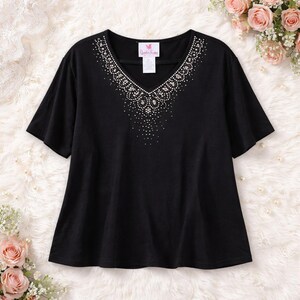 May include: Black short-sleeve top with a V-neckline. The neckline is embellished with a pattern of small, sparkling rhinestones. A small tag with text is visible near the neckline. The top is made of a soft, flowing fabric.