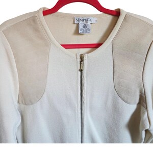 May include: Cream-colored zip-up sweater with beige shoulder patches. The sweater has a round neckline and a gold-colored zipper. The label reads "SEMPLICE". The sweater is hanging on a red hanger.