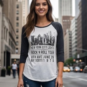 May include: A white raglan t-shirt with three-quarter length dark grey sleeves. The front features a black graphic of a city skyline above the text: NEW YORK CITY LIGHTS, ROCK N ROLL TOUR, 8TH AVE JUNE 26, NY 10011 1 9 7 8.