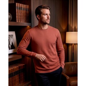 May include: A rust-colored long-sleeve crew neck shirt. The shirt is made of a textured fabric and is paired with dark blue pants. The person is leaning against a wooden bookshelf with leather-bound books.