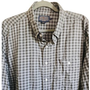 Może przedstawiać: Szara i biała koszula w kratę z kołnierzykiem zapinanym na guziki i kieszenią na piersi. Koszula jest marki Pendleton i wisi na szarym wieszaku.