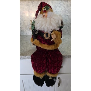 May include: A decorative Santa Claus figure with a burgundy velvet suit, a white beard, and a red hat. The figure has a gold belt buckle and holds a small green tree. The boots are black with gold accents. A festive Christmas decoration.