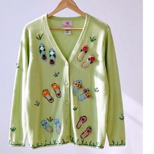 May include: A light green cardigan sweater with a V-neck and button closure. The sweater is decorated with embroidered flip-flop designs in various colors, including red, orange, blue, and pink. Small floral and leaf accents are also present.