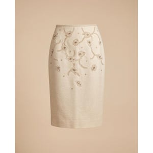 May include: A cream-colored knee-length skirt featuring intricate beadwork in a floral vine pattern. The embellishments are concentrated on the upper portion of the garment, with a clean, solid hemline and a structured waistband at the top.