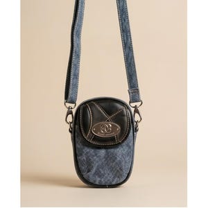 May include: A small crossbody bag featuring a blue denim body with a textured finish and a black leather flap. The flap includes a silver oval metal plaque with the embossed text "Slicil" and "Mccann". It has a matching blue denim shoulder strap.