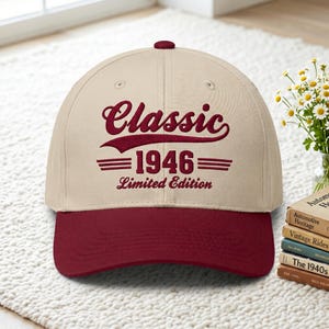 May include: A beige baseball cap with a maroon brim and top button. The cap features the text "Classic 1946 Limited Edition" in maroon script and block letters. The hat is sitting on a white rug.