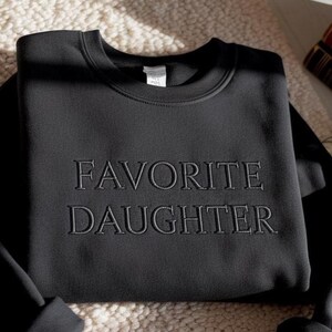 Puede incluir: Sudadera negra con la inscripci&oacute;n bordada "FAVORITE DAUGHTER". La sudadera de cuello redondo est&aacute; doblada. El texto est&aacute; en negrita, en una fuente sans-serif. La tela parece suave.