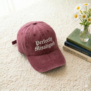 May include: A maroon baseball cap with the text "Perfectly Misaligned" embroidered in white. The cap is made of a soft, textured fabric and has a curved brim. The cap is sitting on a cream-colored carpet.