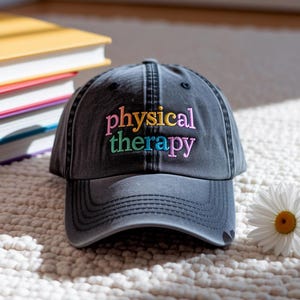 May include: A dark gray baseball cap with the words "physical therapy" embroidered in colorful thread. The hat has a curved brim and a small heart detail. A stack of books and a daisy are in the background.