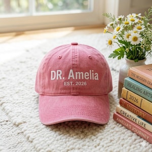 May include: A dusty rose baseball cap with the text "DR. Amelia EST. 2026" embroidered in white. The cap is sitting on a white rug. A vase of white flowers and a stack of books are in the background.