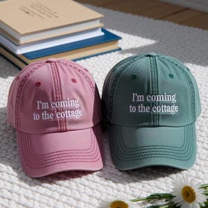 May include: Two baseball caps, one dusty rose and one teal, with the text "I'm coming to the cottage" embroidered in white. The caps are on a white, textured surface, with a stack of books in the background.