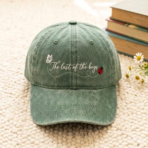 May include: A green baseball cap with the embroidered text "The last of the bugs." The design includes a white butterfly and a red ladybug. The cap is made of a soft, textured fabric.