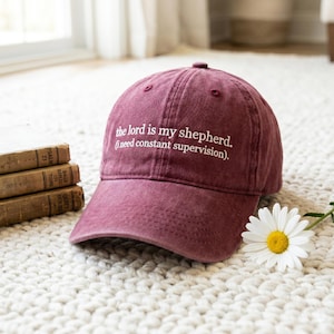 May include: A maroon baseball cap with the text "the lord is my shepherd. (i need constant supervision)" in white. The cap is sitting on a white rug next to a daisy and a stack of books.