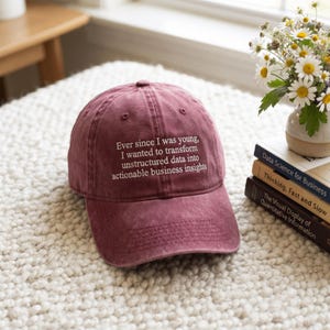 May include: A maroon baseball cap with white embroidered text that reads: Ever since I was young, I wanted to transform unstructured data into actionable business insights. The cap features a curved brim and a structured crown with ventilation eyelets.