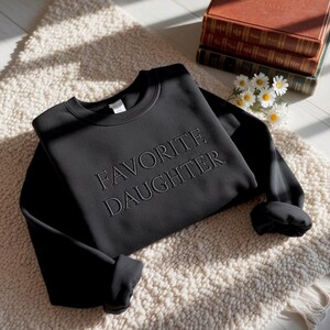 Puede incluir: Sudadera negra con el texto bordado "FAVORITE DAUGHTER". La sudadera de cuello redondo se muestra sobre una alfombra color crema, con una pila de libros y margaritas en el fondo.
