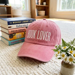 May include: A pink baseball cap with the text "BOOK LOVER" embroidered in white. The cap is next to a stack of books with titles including "The Midnight Library" and "Educated". A small vase of daisies is also in the image.