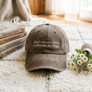 May include: Brown baseball cap with the text "i don't rise and shine, i caffeinate and hope." The hat is sitting on a white rug next to a stack of books and a vase of flowers. The hat is a neutral color.