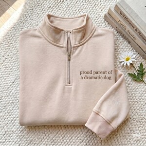 May include: A beige quarter-zip sweatshirt featuring a stand-up collar and long sleeves with ribbed cuffs. The text "proud parent of a dramatic dog" is embroidered in brown on the upper left chest area of the garment.