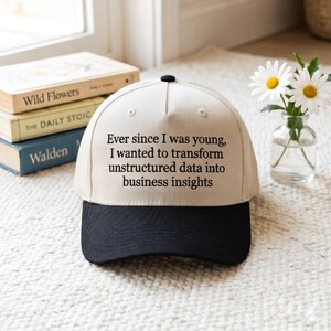 May include: A two-tone baseball cap with a cream-colored crown and a black brim. The front of the crown features the black printed text: Ever since I was young, I wanted to transform unstructured data into business insights. The cap has a button top.