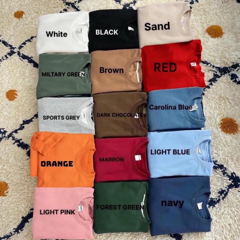 Puede incluir: Una colecci&oacute;n de sudaderas de cuello redondo dobladas en varios colores, incluyendo blanco, negro, arena, verde militar, marr&oacute;n, rojo, gris deportivo, chocolate oscuro, naranja, granate, rosa claro, verde bosque, azul claro, azul Carolina y marino. Cada camisa tiene una etiqueta de color.