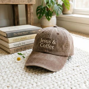 May include: A brown baseball cap with the embroidered text "Jesus & Coffee" in white. The cap is made of a soft, worn material and has a curved brim. The cap is sitting on a white, textured surface.