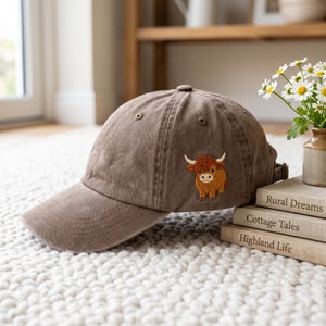 May include: A brown baseball cap featuring an embroidered graphic of a small, fluffy Highland cow on the side. The cap has a curved brim, stitched eyelets on the crown, and an adjustable strap at the back, showcasing a simple and functional design.