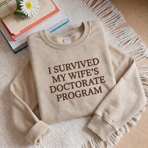 May include: A beige sweatshirt with the embroidered text "I SURVIVED MY WIFE'S DOCTORATE PROGRAM" in brown. The sweatshirt is folded on a white rug with a stack of books and a daisy.