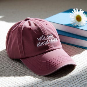 May include: A dusty rose baseball cap with the embroidered text "will yap about books" in white. The cap is on a white textured surface, with blue books and a daisy in the background.