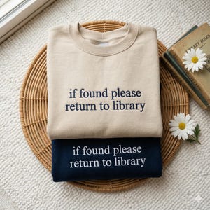 May include: Two sweatshirts, one beige and one navy blue, stacked on a woven tray. Each sweatshirt has the embroidered text "if found please return to library" in a contrasting color. A book and daisies are in the background.