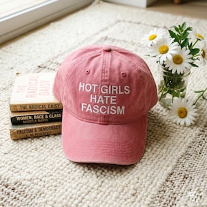 May include: A dusty rose baseball cap with the text "HOT GIRLS HATE FASCISM" in white. The hat is on a textured rug next to a stack of books and a small vase of daisies.