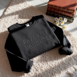 Puede incluir: Sudadera negra con el texto bordado "FAVORITE DAUGHTER". La sudadera de cuello redondo se muestra sobre una alfombra color crema, con una pila de libros y margaritas en el fondo.