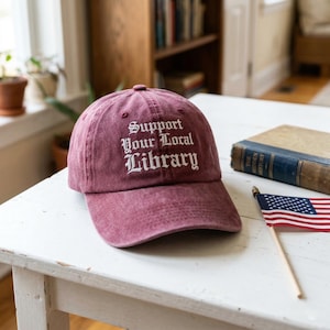 May include: A maroon baseball cap with the text "Support Your Local Library" in white script. The cap is on a white table next to a small American flag and a vintage book.