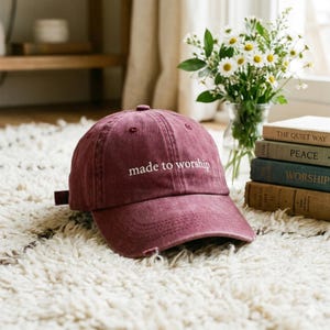 May include: A maroon baseball cap with the text "made to worship" embroidered in white. The cap is distressed, with a slightly worn appearance. The background includes a fluffy white rug, books, and a vase of flowers.