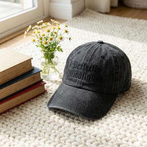 May include: A black, distressed baseball cap with the embroidered text "Perfectly Misaligned" in a gothic font. The cap is sitting on a cream-colored, textured rug. A small vase of daisies and a stack of books are in the background.