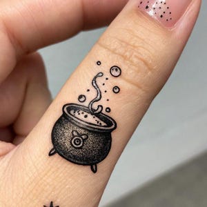 Könnte beinhalten: Schwarz-weiß Tattoo eines brodelnden Kessels mit Dampf und Blasen. Unter dem Kessel befinden sich ein Stern und ein Halbmond mit einem Stern. Das Tattoo befindet sich auf einem Finger.